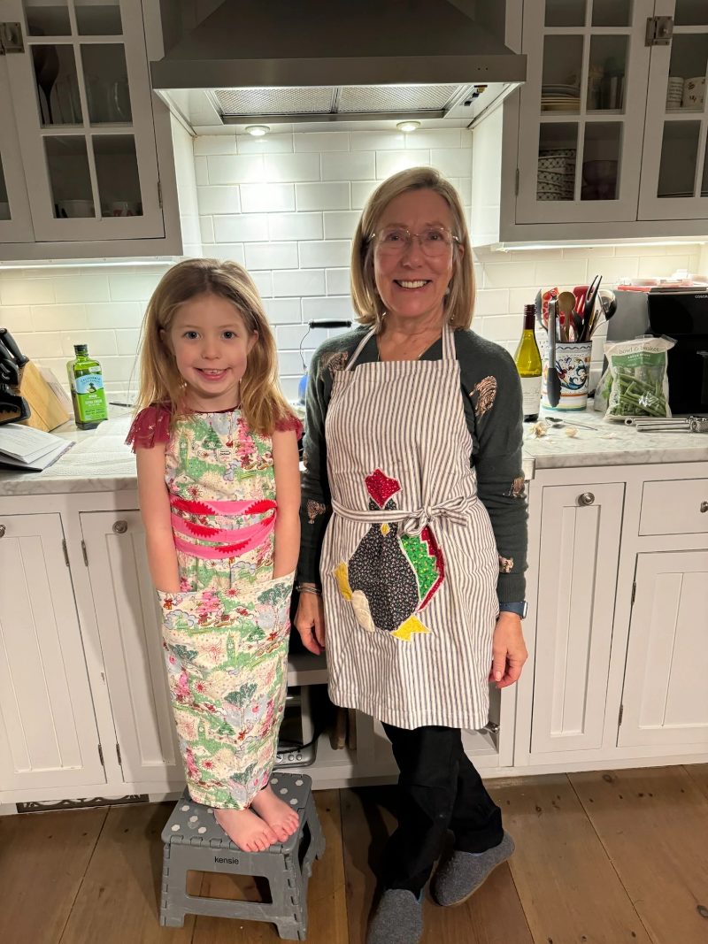 Grandmother in her apron and her granddaughter, up on a step stool, cooking together in the kitchen